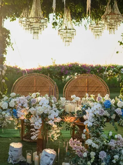 A romantic sweetheart table for an outdoor garden wedding, set under a canopy of hanging chandeliers and surrounded by lush blue and white flowers.