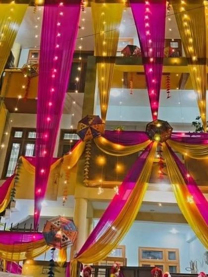 A stunning night-time house decoration with yellow and pink drapes, fairy lights, and hanging Rajasthani umbrellas. The entire house glows with festive energy.