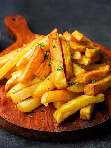 A pile of classic salted french fries on a wooden board with a side of ketchup. The dark background and simple styling make this a timeless and appealing shot.