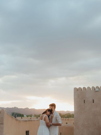 A serene moment overlooking a historic fort in Muscat, Oman. The couple embraces as the sun sets behind the mountains, casting a warm glow over the ancient landscape.