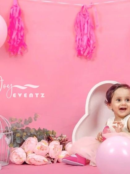 A delightful pink-themed first birthday cake smash. The setup includes a heart-shaped prop, pink balloons, and floral arrangements, capturing the happy expressions of the birthday girl as she celebrates her special day.