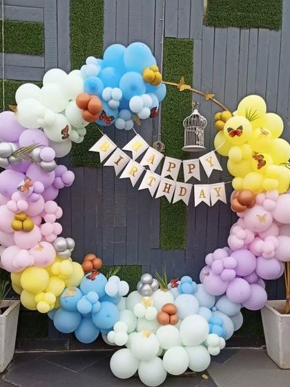 A whimsical outdoor birthday setup with a circular balloon arch in pastel colors. The arch is decorated with a 'Happy Birthday' banner and small butterfly accents.