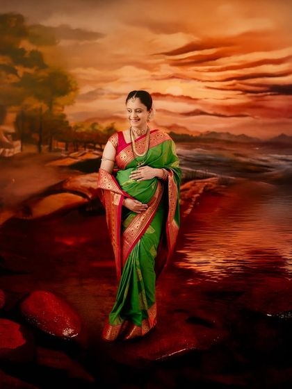 A beautiful portrait set against a painted sunset backdrop. The rich colors of the sky and water complement the vibrant green and red of the traditional saree.