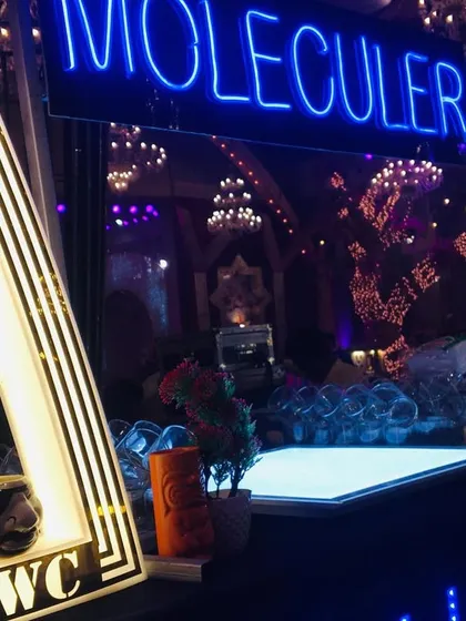 A close-up of the 'Moleculer' bar setup at a grand event. The custom 'TWC' light and the deep blue neon sign create a premium and inviting look.
