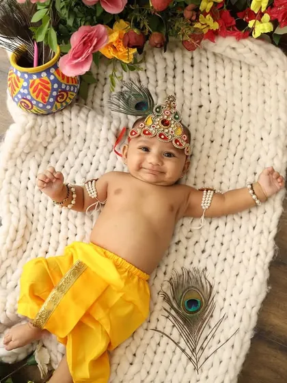 A happy baby Krishna lying on a soft blanket, surrounded by peacock feathers. His joyful expression is simply contagious.
