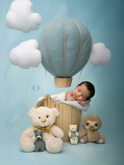 A tiny newborn sleeps in a basket designed as a hot air balloon, floating among plush clouds and teddy bears. This whimsical and creative setup turns a simple portrait into a delightful adventure scene.