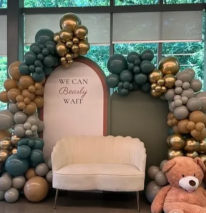 An elegant "We Can Bearly Wait" baby shower decor in a sophisticated palette of sage green, beige, and chrome gold. The setup includes a comfortable scalloped sofa for the mom-to-be, a giant teddy bear, and lush balloon arches for a luxurious feel.