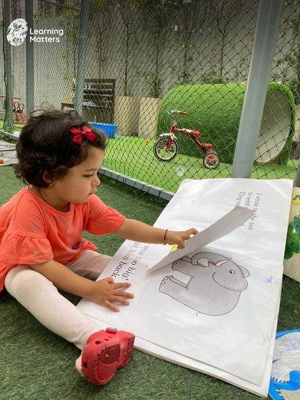 A love of books is one of the greatest gifts we can give a child. Here, a toddler explores a large picture book in our outdoor area, connecting the stories on the page with the world around her.