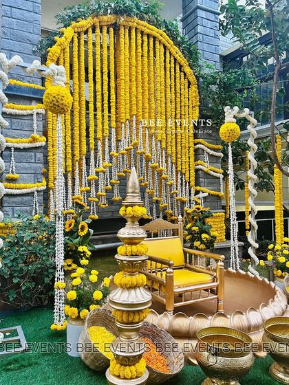 A beautiful and elaborate Haldi setup. The seating area is framed by an arch of marigold garlands, with traditional brass props and a lotus-shaped 'urli' for the ceremony, creating a truly grand and auspicious look.