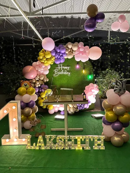 A vibrant first birthday decoration with a jungle-grass backdrop and a colorful mix of pink, purple, and gold balloons.