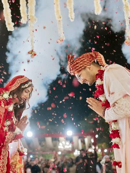 A moment of reverence during the Varmala ceremony. The couple is framed by dramatic smoke and a shower of petals, with hanging tuberoses and chandeliers above.