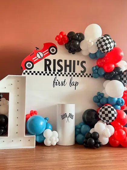 Ready, set, go! This is a high-energy racing car theme for "Rishi's first lap," featuring a large light-up number '1' and a custom backdrop. The theme is driven home with a dynamic balloon arch in red, black, white, and blue, including fun checkered-flag pattern balloons.