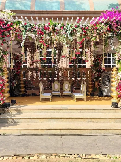 A beautiful outdoor mandap setup under a pergola structure, lavishly decorated with pink and white flowers and hanging floral strings for a romantic daytime ceremony.