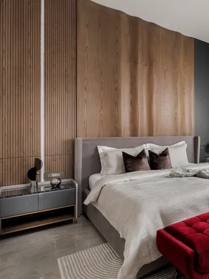 We designed this son's bedroom with a cool and cozy mix of materials. A dramatic, undulating wooden headboard is the star of the room, complemented by a grey upholstered bed and a bold pop of color from the red velvet ottoman.