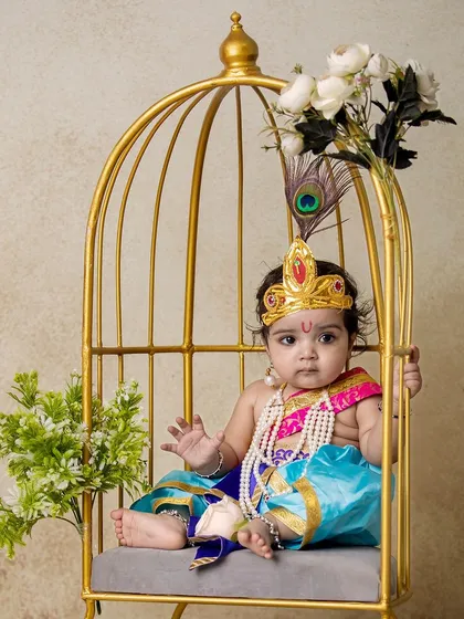 A baby Krishna sitting inside a golden birdcage prop, decorated with flowers. This is one of my unique props that adds a touch of fantasy to the shoot.