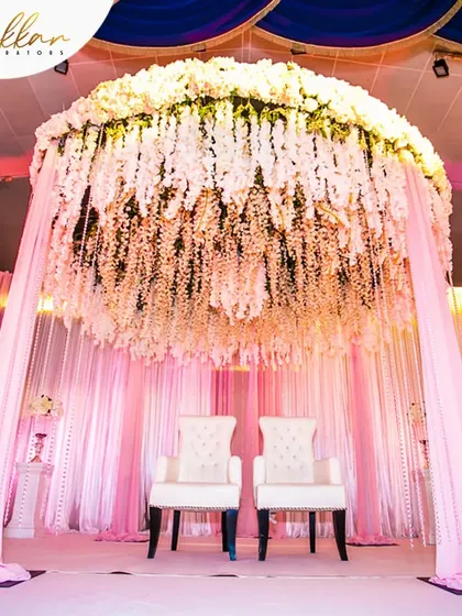 A stunning mandap or stage design with a canopy made entirely of hanging white flowers. The soft pink lighting creates a dreamy and ethereal ambiance.