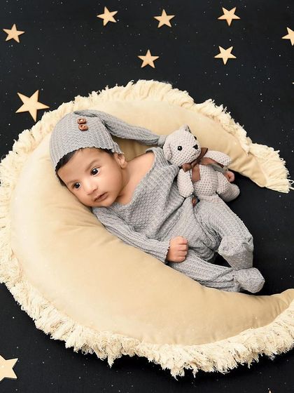 To the moon and back. This little one, dressed in a cozy grey outfit, rests on a crescent moon against a starry night sky.