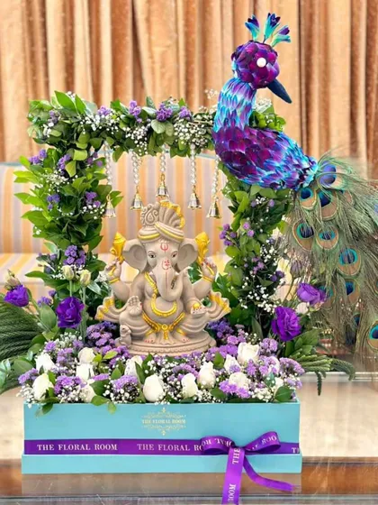 A truly unique and spiritual floral arrangement featuring a Ganesha idol. The idol is surrounded by a beautiful arch of fresh purple and white flowers, with a decorative peacock accent, all set in a custom gift box.