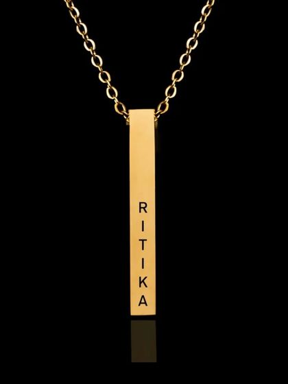 A studio shot of a gold bar pendant engraved with the name 'Ritika'. The black background makes the polished gold and crisp engraving stand out, highlighting the quality of the piece.