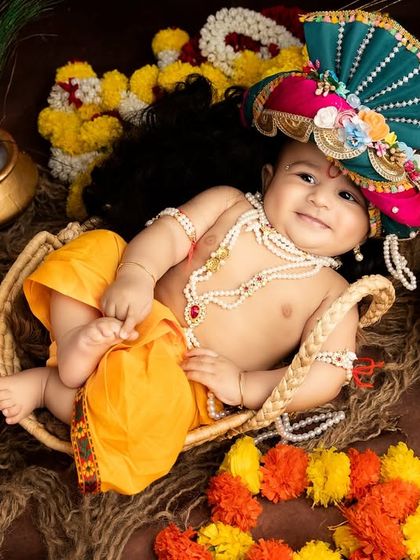 A joyful baby Krishna lying in a basket, surrounded by colorful flowers and pots. His smile truly lights up the photo.