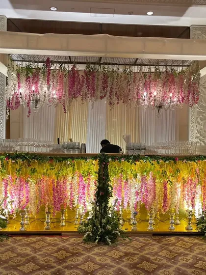 An elaborate and beautiful bar setup for a wedding. The bar front is decorated with hanging wisteria flowers, and the structure is topped with a matching floral arrangement.