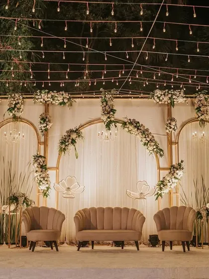 A beautiful and sophisticated stage design for a reception. The backdrop features gold arches, sheer white drapes, and delicate floral arrangements, creating a look of timeless elegance.