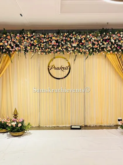 A beautiful engagement stage with a custom name sign set against a backdrop of golden and cream drapes. The setup is framed by lush, colorful floral arrangements on the top and sides.
