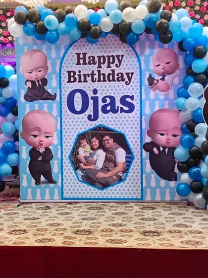 A full view of the Boss Baby stage setup, featuring a personalized backdrop with a family photo and multiple character poses. The blue, black, and white balloon garland completes the look.