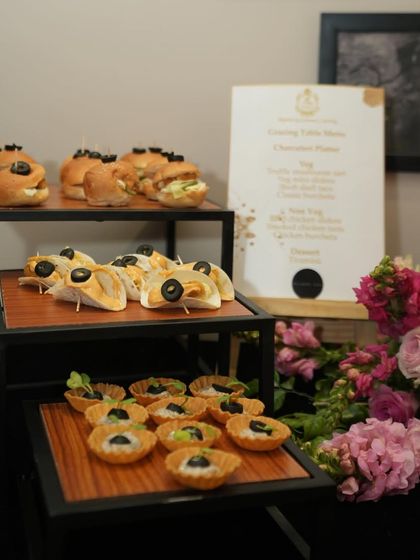 A three-tiered stand showcasing a variety of savory bites, including mini tacos, tarts, and sliders. The custom menu for the Bhawna Rao event is visible in the background.