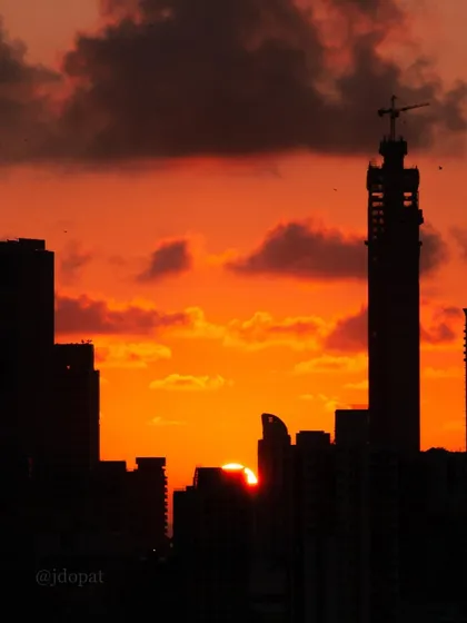 The sun is partially hidden behind a silhouetted building under construction, creating a dramatic orange sky over Mumbai.