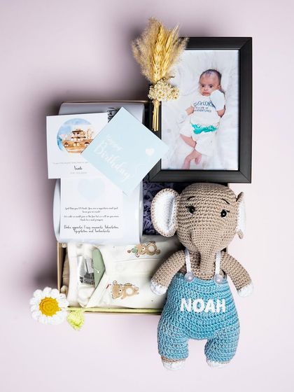 A sweet hamper curated for a newborn baby named Noah. It features a personalized crochet elephant toy, a baby onesie, and a framed photo of the little one.