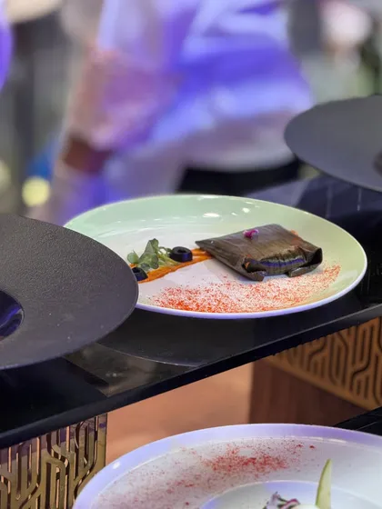 A dish wrapped in a banana leaf, presented on a plate with a colourful spice dusting. The presentation hints at the traditional flavours held within a modern aesthetic.
