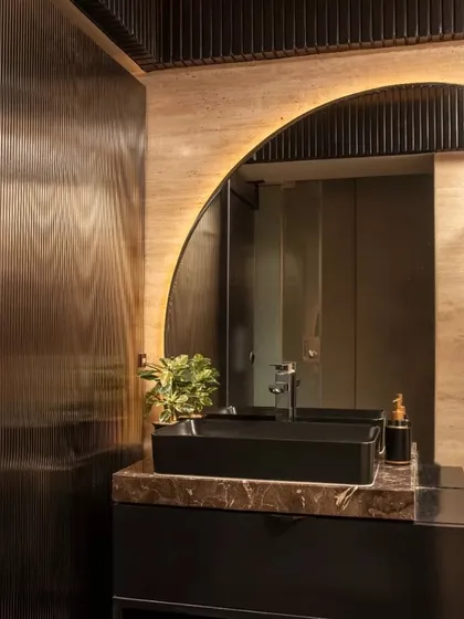 A powder room with a dark, moody aesthetic, featuring a black basin, dark wood paneling, and a large, arch-shaped backlit mirror.
