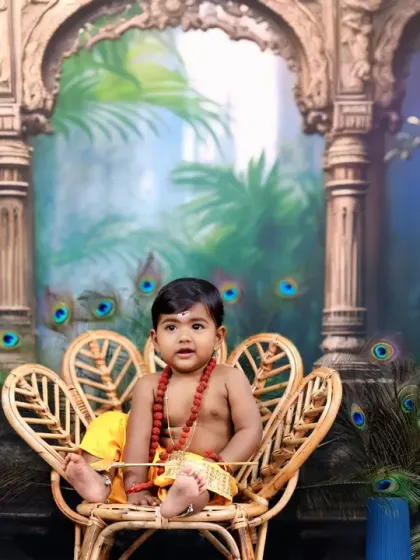Dressed as little Muruga, this child sits in a peacock-shaped wicker chair, surrounded by a palatial backdrop and real peacock feathers, creating a majestic scene.