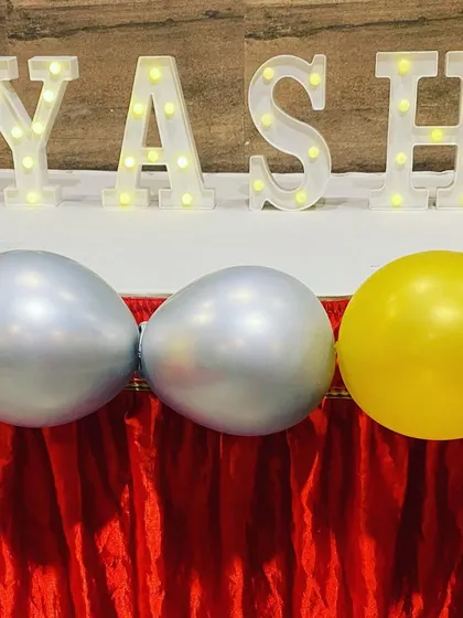 Light-up marquee letters spelling "YASH" on a table decorated with balloons for a first birthday party.