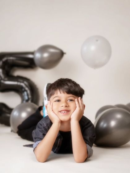A playful and relaxed pose from a fifth birthday shoot. Lying amongst the balloons, he looks directly at the camera with a sweet, contemplative smile. This candid style of portrait captures the quiet, happy moments of a celebration.