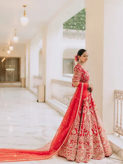A portrait of the bride in a classic red lehenga, looking out a window. The soft, natural light and elegant pose create a feeling of timeless grace.
