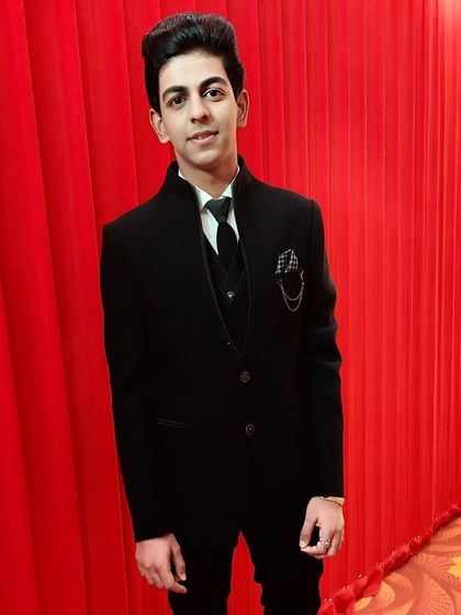 This young man is styled for a wedding party in a sharp black suit. The hair is styled for volume and the makeup is minimal, giving him a youthful yet sophisticated look.
