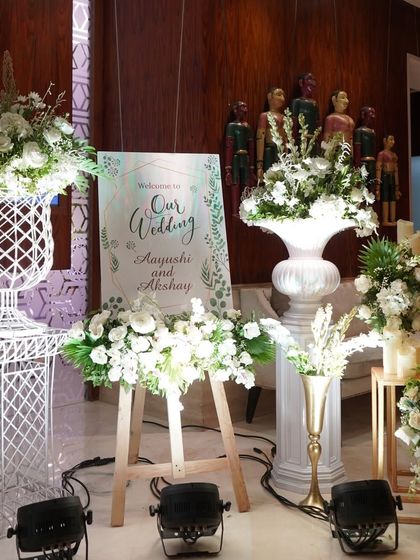 A clean and classic welcome setup. The easel with the couple's welcome sign is surrounded by beautiful arrangements of white flowers in classic urns, creating a timeless and elegant first impression.