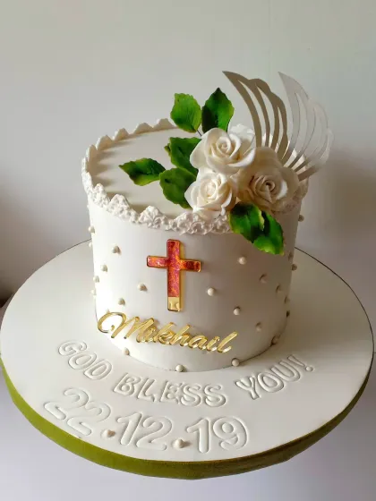 An elegant and classic First Communion cake. The clean white design is decorated with a golden cross, sugar pearls, and a spray of white sugar roses. The message "God Bless You" adds a personal touch to the celebration.