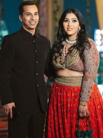 A stylish couple in formal Indian wear at a large family celebration or wedding reception.