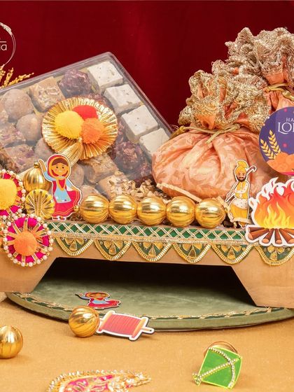 This festive Lohri hamper is arranged on a decorative stand and includes a box of assorted mithai, golden laddus, and potlis, all adorned with fun, colourful cutouts.