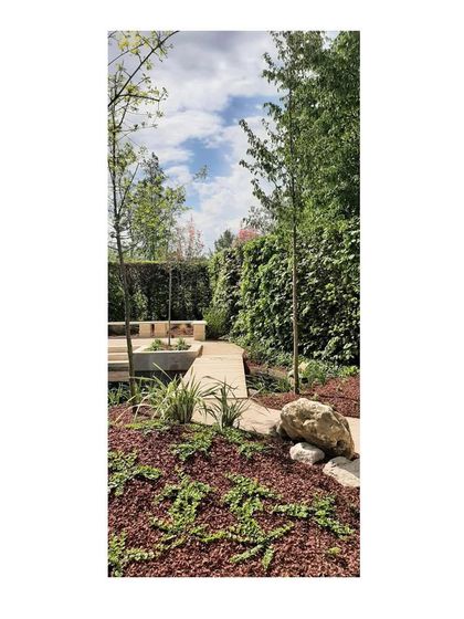 The pathway curves gently, guiding the visitor's journey through the garden. The use of natural stone and red bark mulch adds texture and color, complementing the green foliage.