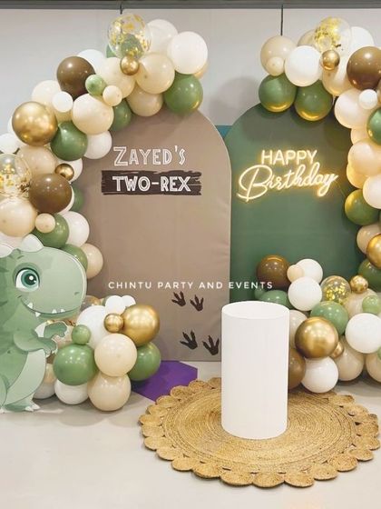 A 'Two-Rex' dinosaur-themed second birthday party. This cute setup features a dinosaur cutout, custom 'Zayed's Two-Rex' backdrop, and an organic balloon arch in earthy green, brown, and gold tones.