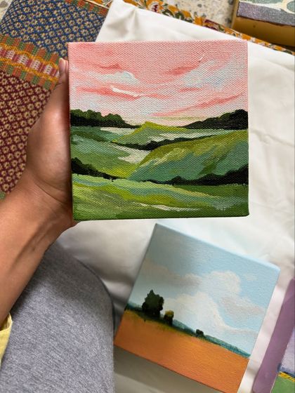 Holding a miniature painting of rolling green hills under a soft pink sky. This demonstrates the personal, handheld scale of these small but 'scenery-ous' artworks.