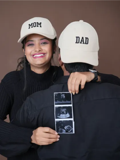 A fun and creative announcement shot. The couple wears "Mom" and "Dad" hats while holding up their baby's first ultrasound pictures, sharing their joy in a playful way.