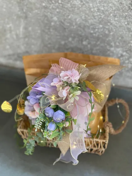 A single wicker basket hamper illuminated by fairy lights and topped with a beautiful arrangement of lavender and pink flowers. This image captures the magical and whimsical quality of our gifts.