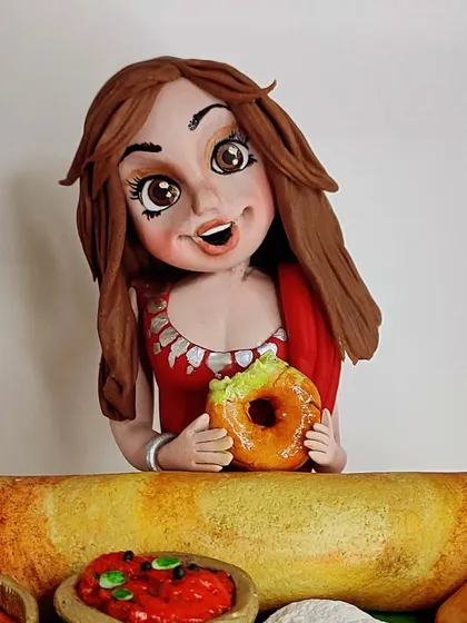 A close-up of the South Indian food cake topper. The figurine is holding a tiny, edible medu vada, capturing a moment of pure foodie joy.