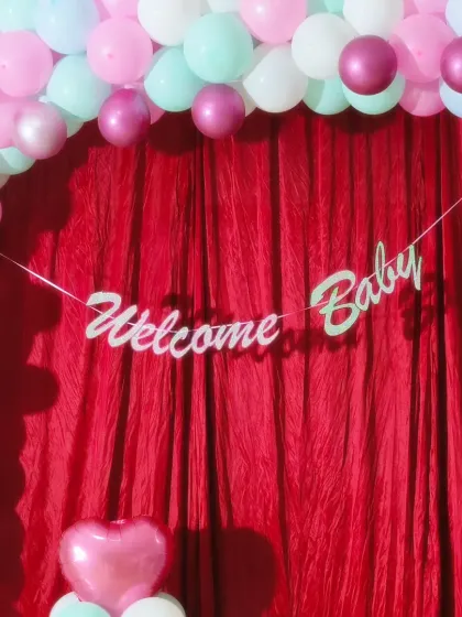 A "Welcome Baby" banner against a red curtain, framed by a pastel balloon arch. This is a great option for a simple stage setup at home.
