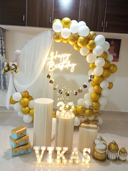 A personalized birthday setup featuring a half-ring balloon arch in gold and white against a sheer curtain. I also included a photo banner with pictures of the birthday person, light-up marquee letters, and decorative props.
